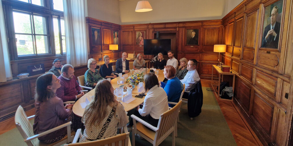 Människor sitter runt konferensbord i träpanelklädd mötesrum.