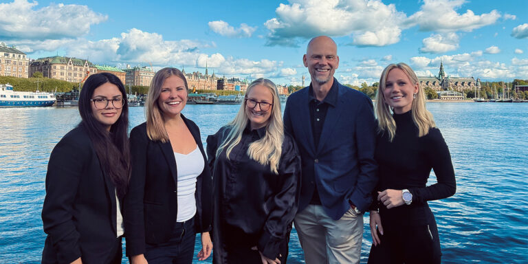 Fem personer poserar framför sjö och stadssilhuett.