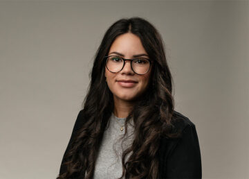 Kvinna står framför neutral bakgrund, bär glasögon och leende.