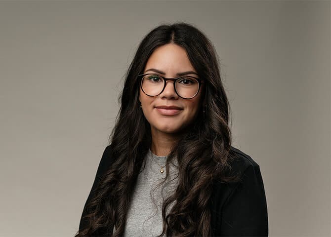 Kvinna står framför neutral bakgrund, bär glasögon och leende.