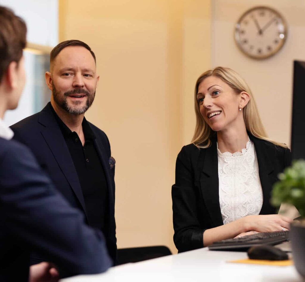 Personer samtalar leende vid skrivbord på kontor. Klocka bakom.