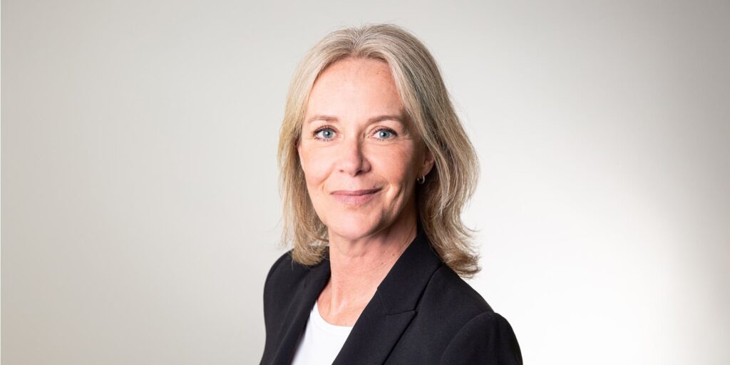 Kvinna i kostymjacka ler framför ljus bakgrund.