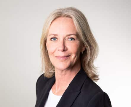 Kvinna i kostymjacka ler framför ljus bakgrund.