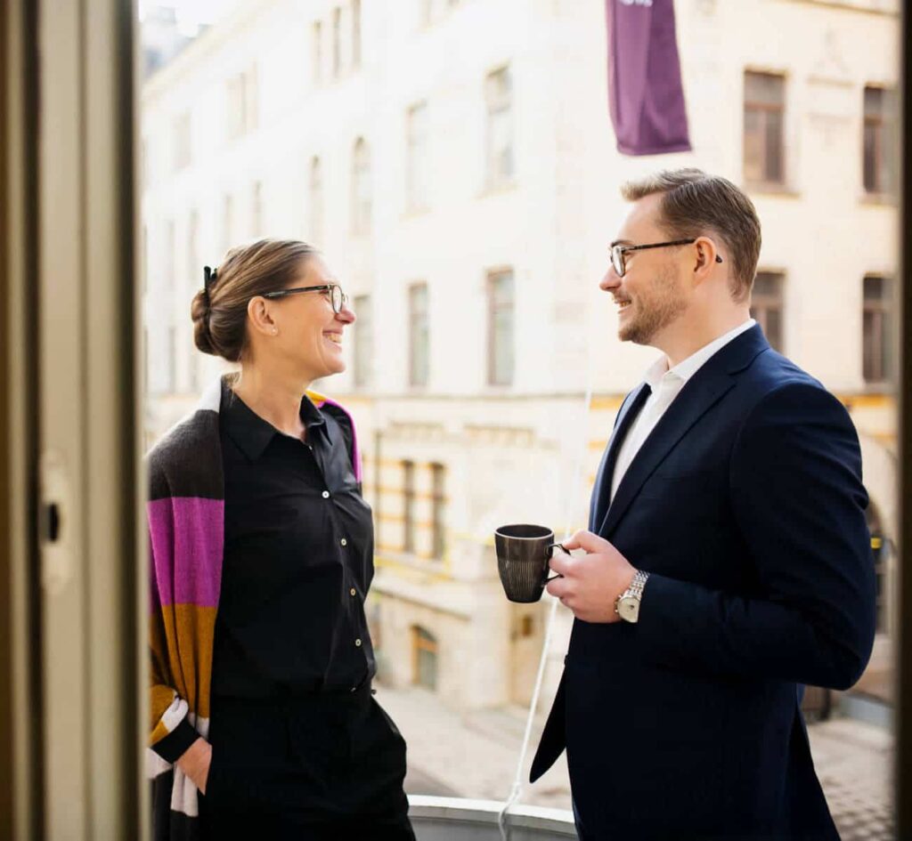 Två personer pratar vänligt på en balkong med byggnader i bakgrunden.