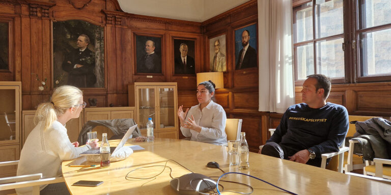 Tre personer diskuterar vid bord i träpanelrum.