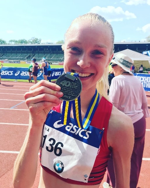 Kvinna håller medalj leende efter lopp på idrottsarena.