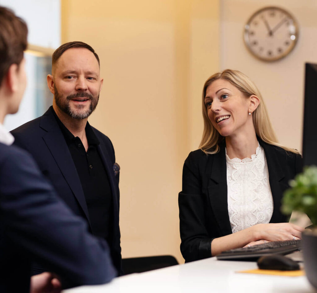 Personer samtalar glatt på kontor framför dator och klocka.