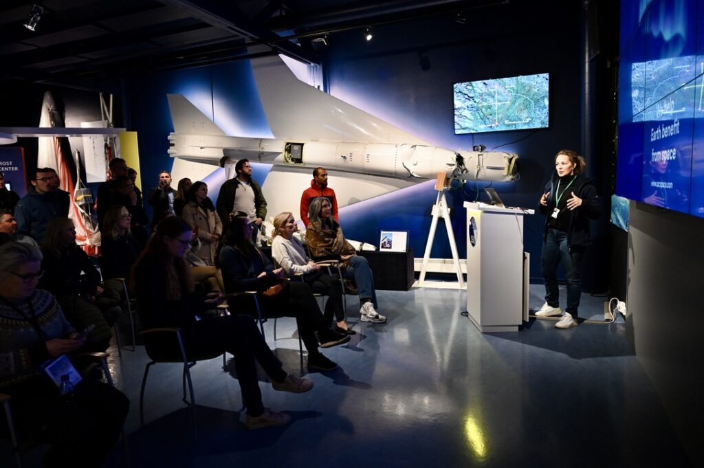 Publik föreläsning om rymden framför flygplansutställning, åhörarna lyssnar.