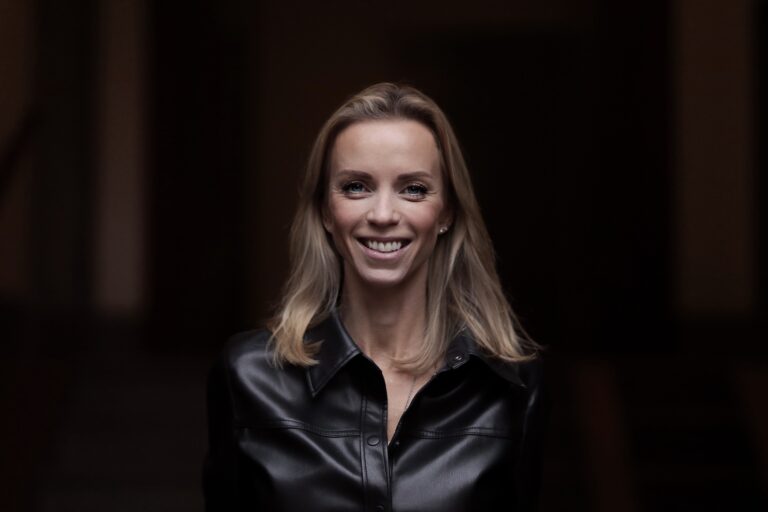 Leende kvinna står i mörk jacka, inomhus bakgrund.