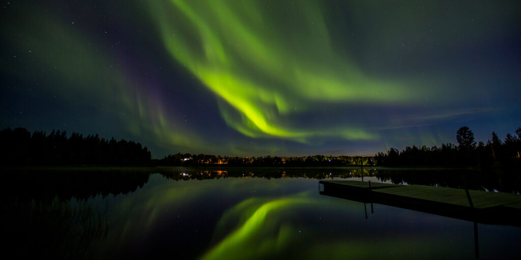 Norrsken reflekteras över lugn sjö med mörk skog.