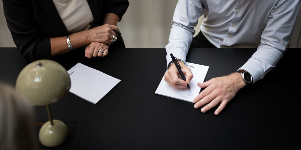 Hand signerar dokument på bord, bredvid observant person.