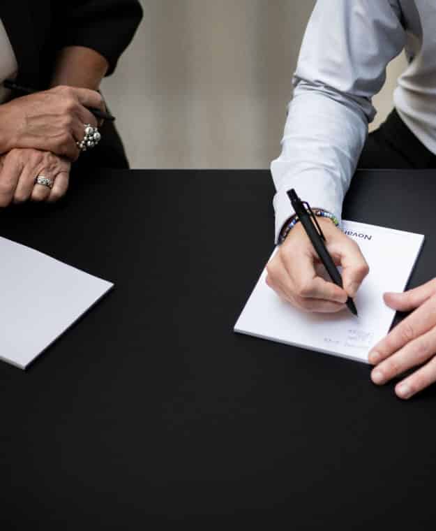 Hand signerar dokument på bord, bredvid observant person.