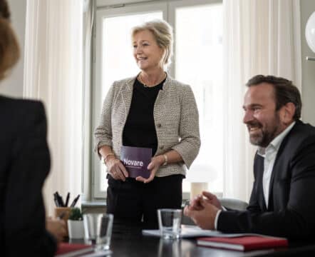 Kvinna håller "Novare"-kort, samtalar vid konferensbord med kollegor.