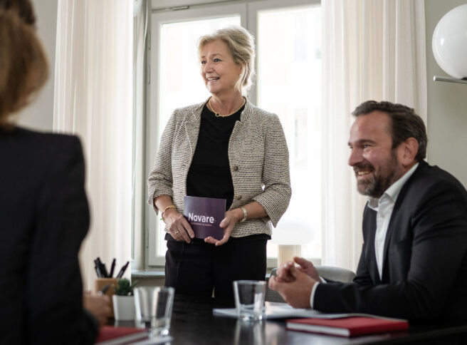 Kvinna håller "Novare"-kort, samtalar vid konferensbord med kollegor.