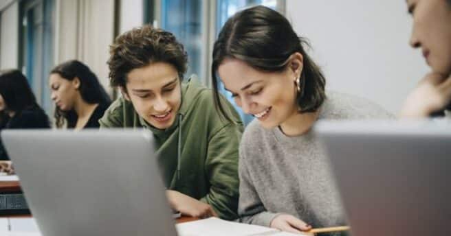 Studenter studerar tillsammans vid laptops i ett klassrum.