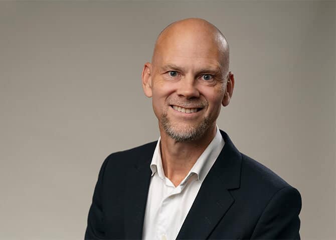 Man ler klädd i kostym mot grå bakgrund.