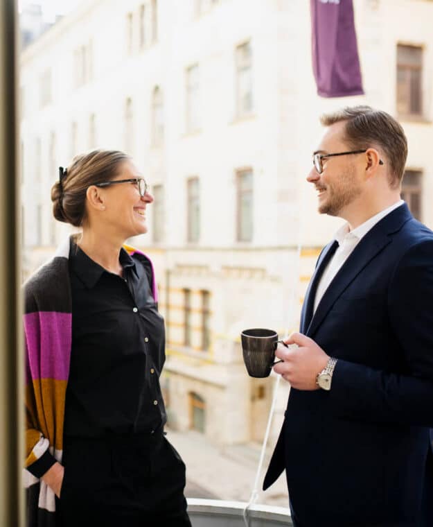 Två personer samtalar på balkong med kaffekopp, stadsbakgrund.