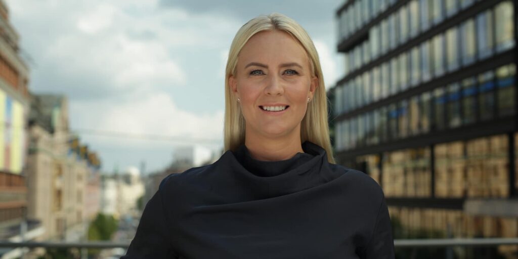 Kvinna ler utomhus med en byggnad i bakgrunden.