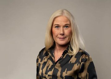 Kvinna i mönstrad blus tittar mot kamera, neutral bakgrund.