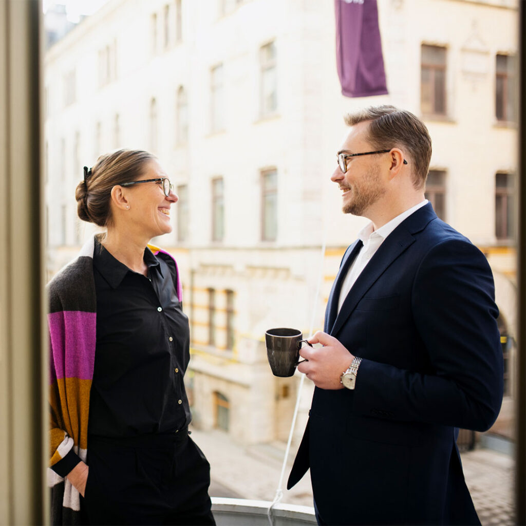 En man med en kopp i handen står och pratar med en kvinna på en balkong.