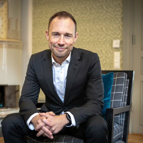 En man i svart kostym sitter i en fotölj och ler mot kameran.