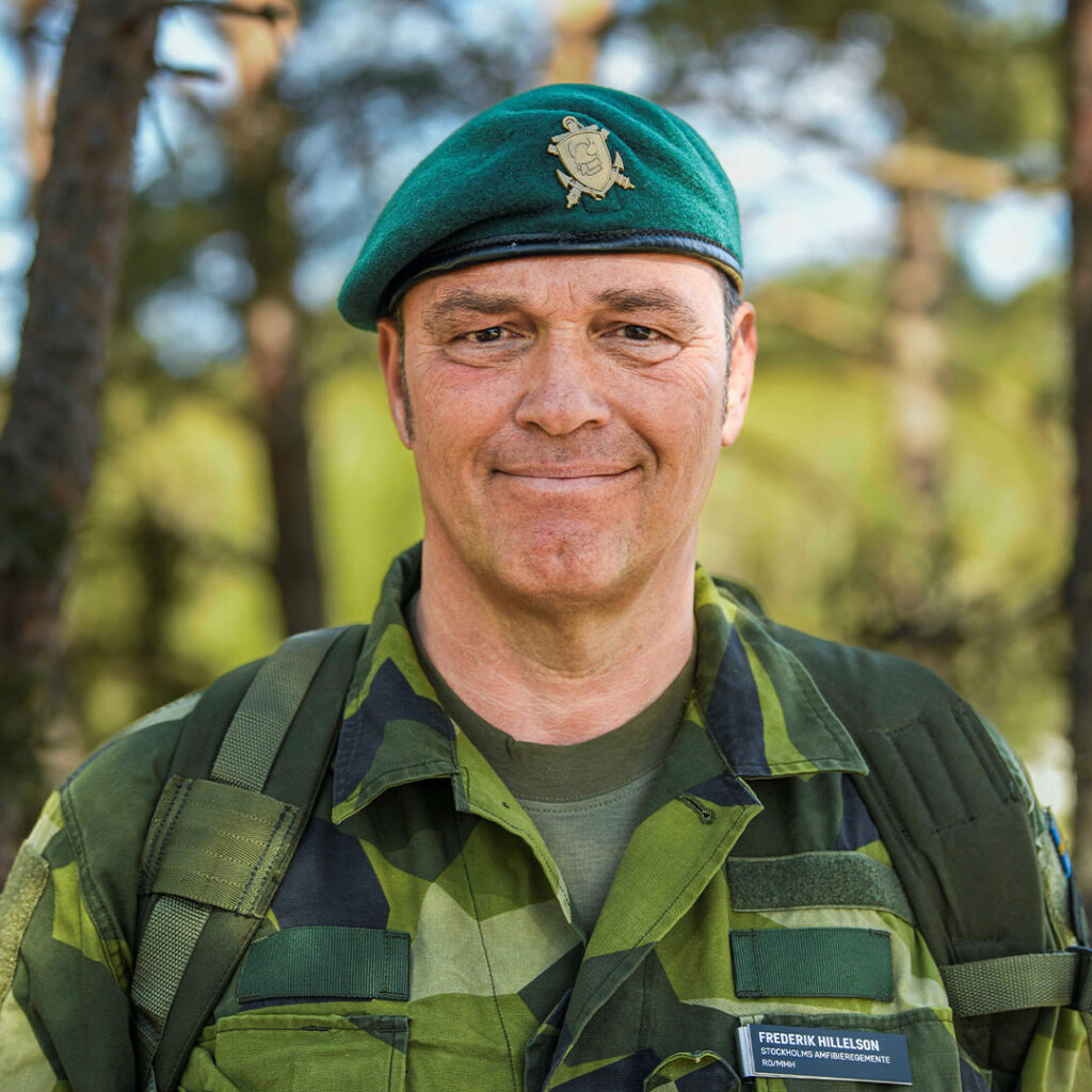 En man i skogen, iklädd uniform som ler mot kameran.