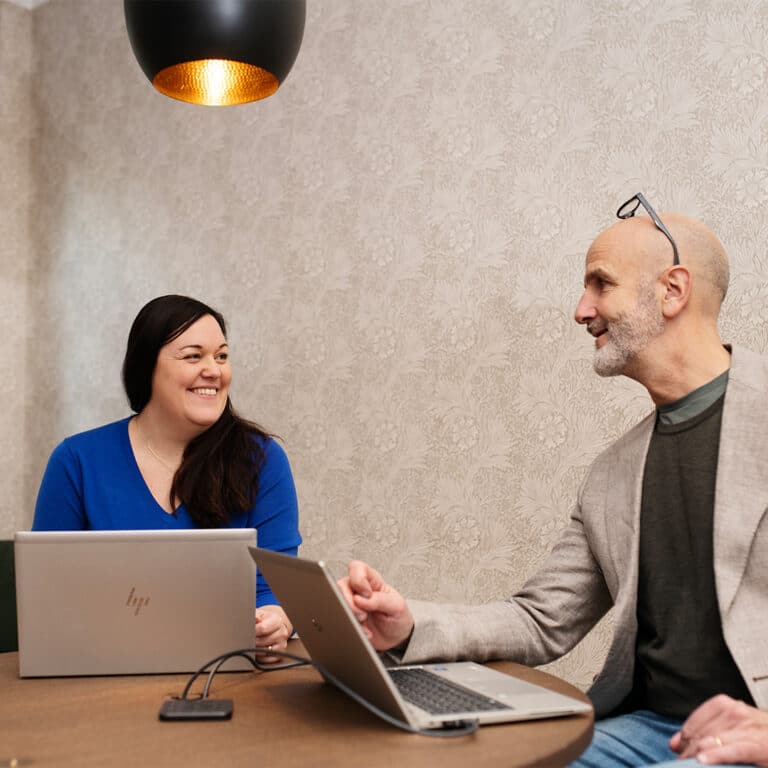 En kvinna och en man sitter med varsina datorer och ler mot varandra.