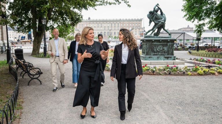 Två kvinnor går och samtalar. Tre personer syns i bakgrunden.