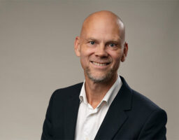 Man ler klädd i kostym mot grå bakgrund.